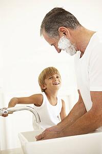Grandson Watching Grandfather Shaving In Bathroom Mirror