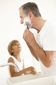 Grandson Watching Grandfather Shaving In Bathroom Mirror