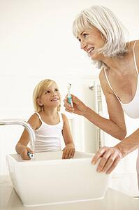 Grandmother Brushing Teeth In Bathroom With Granddaughter Watching