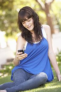 Portrait Of Teenage Girl Sitting In Park Using Mobile Phone