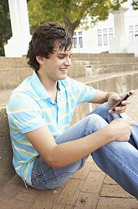 Male Teenage Student Sitting Outside On College Steps Using Mobile Phone