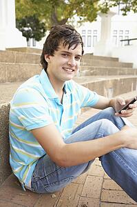 Male Teenage Student Sitting Outside On College Steps Using Mobile Phone