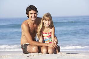 Father And Daughter Wearing Swimwear Sitting On Sandy Beach