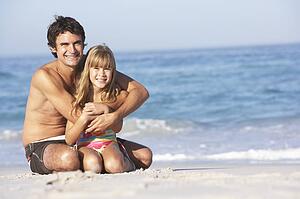 Father And Daughter Wearing Swimwear Sitting On Sandy Beach