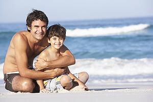 Father And Son Wearing Swimwear Sitting On Sandy Beach