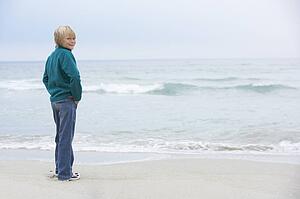 Young Boy On Holiday Standing On Winter Beach Looking Out To Sea
