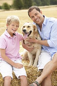 Father And Son Sitting With Dog On Straw Bales In Harvested Field