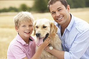 Father And Son Sitting With Dog On Straw Bales In Harvested Field