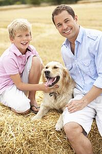 Father And Son Sitting With Dog On Straw Bales In Harvested Field