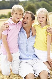 Father And Children Sitting On Straw Bales In Harvested Field