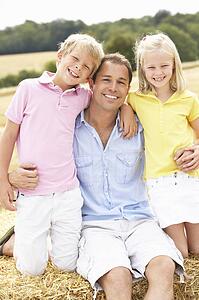 Father And Children Sitting On Straw Bales In Harvested Field