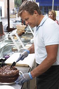 Man Working Behind Counter In Café Slicing Cake