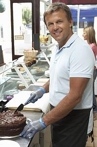 Man Working Behind Counter In Café Slicing Cake