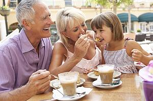 Grandparents With Granddaughter Enjoying Coffee And Cake In Café