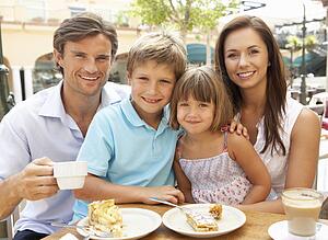Young Family Enjoying Cup Of Coffee And Cake In Café Together