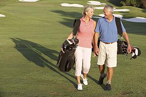 Senior Couple Walking Along Golf Course Carrying Bags