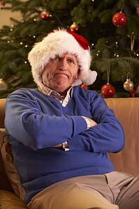 Senior Man Dressed As Father Christmas Looking Grumpy In Front Of Christmas Tree