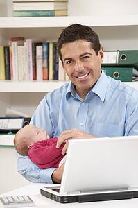 Father With Newborn Baby Working From Home Using Laptop