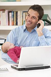 Father With Newborn Baby Working From Home Using Laptop