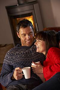 Middle Aged Couple Sitting On Sofa By Cosy Log Fire With Hot Drink