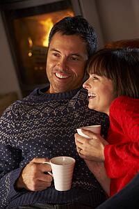 Middle Aged Couple Sitting On Sofa By Cosy Log Fire With Hot Drink