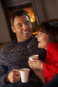 Middle Aged Couple Sitting On Sofa By Cosy Log Fire With Hot Drink