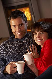 Middle Aged Couple Sitting On Sofa By Cosy Log Fire With Hot Drink