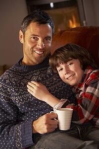 Father And Son Relaxing With Hot Drink Watching TV By Cosy Log Fire