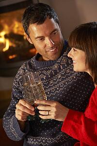 Middle Aged Couple Sitting Sofa By Cosy Log Fire With Glass Of Champagne