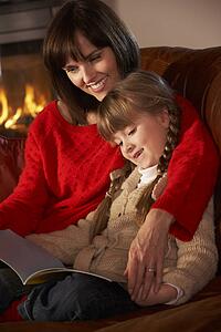Mother And Daughter Sitting On Sofa And Reading Book By Cosy Log Fire