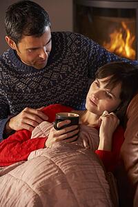 Husband Nursing Sick Wife With Cold Resting On Sofa By Cosy Log Fire