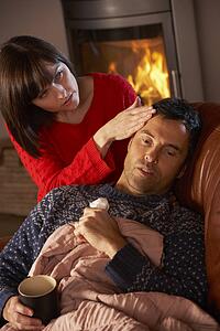 Wife Nursing Sick Husband With Cold Resting On Sofa By Cosy Log Fire