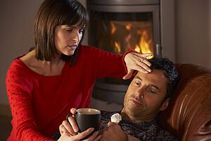Wife Nursing Sick Husband With Cold Resting On Sofa By Cosy Log Fire