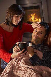 Wife Nursing Sick Husband With Cold Resting On Sofa By Cosy Log Fire