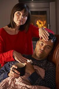 Wife Nursing Sick Husband With Cold Resting On Sofa By Cosy Log Fire