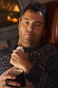 Sick Man With Cold Resting On Sofa By Cosy Log Fire
