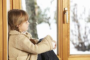 Young Girl Sitting On Window Ledge Looking At Snowy View