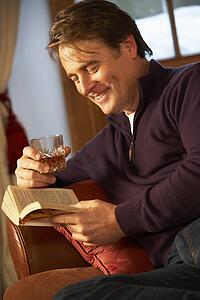 Middle Aged Man Relaxing With Book Sitting On Sofa Drinking Whisky