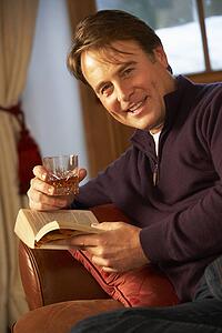 Middle Aged Man Relaxing With Book Sitting On Sofa Drinking Whisky