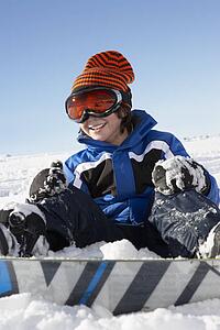 Young Boy Sitting In Snow With Snowboard