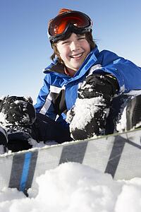 Young Boy Sitting In Snow With Snowboard