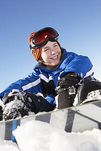 Young Boy Sitting In Snow With Snowboard