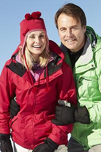 Couple Standing In Snow Wearing Warm Clothes On Ski Holiday In Mountains