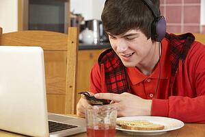 Teenage Boy Using Laptop And Listening To MP3 Player Whilst Eating Breakfast