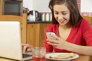 Teenage Girl Using Laptop And Mobile Phone Whilst Eating Breakfast