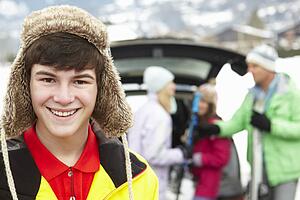 Teenage Boy Smiling At Camera Whilst Family Load Skis In Boot Of Car