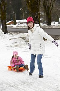 Mother Pulling Daughter On Sledge Along Snowy Street In Ski Resort